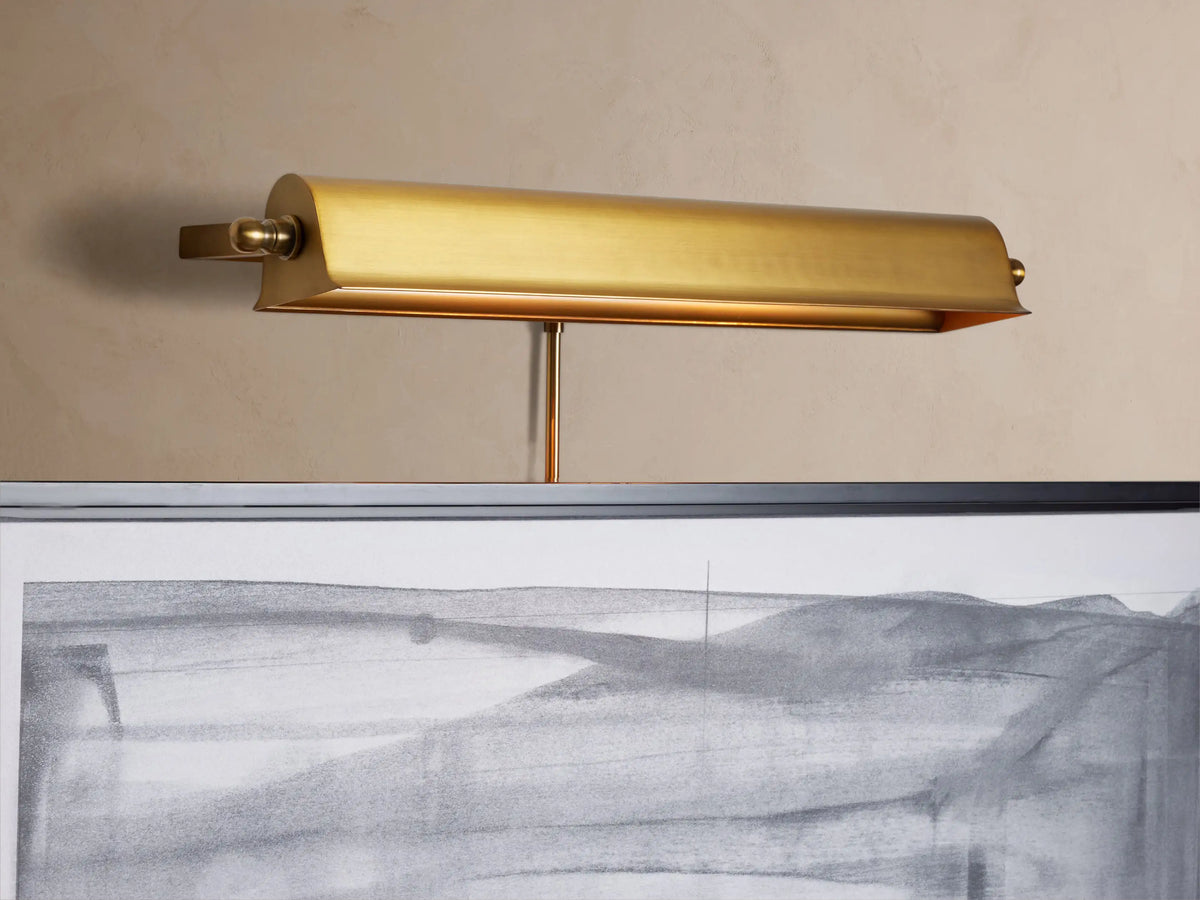 Angled close-up of an antiqued brass Tremont Picture Light Sconce, glowing warmly, mounted on a light beige wall.
