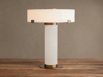 Chayla Table Lamp with glowing white alabaster shade, cylindrical alabaster base, and antique brass accents on a wooden table.