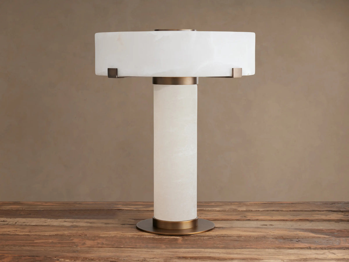 Chayla Table Lamp, white alabaster cylindrical base & drum shade, antique brass accents, on a rustic wooden table.