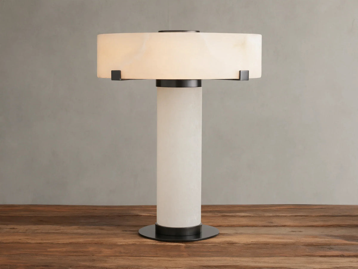 Chayla Table Lamp with cylindrical alabaster base, wide circular alabaster shade, and dark brass accents on a wooden surface.