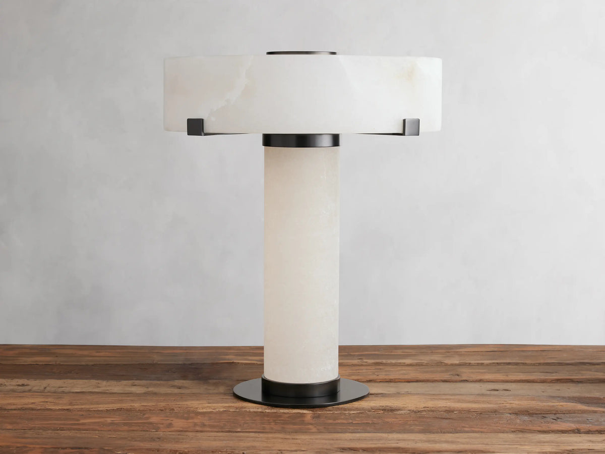Chayla Table Lamp with cylindrical alabaster base, wide circular alabaster shade, and dark brass accents on a wooden surface.
