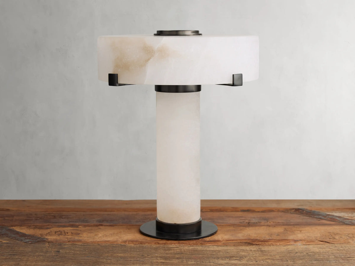 Chayla Table Lamp with cylindrical alabaster base & wide alabaster shade, supported by dark bronze, glowing on wood.