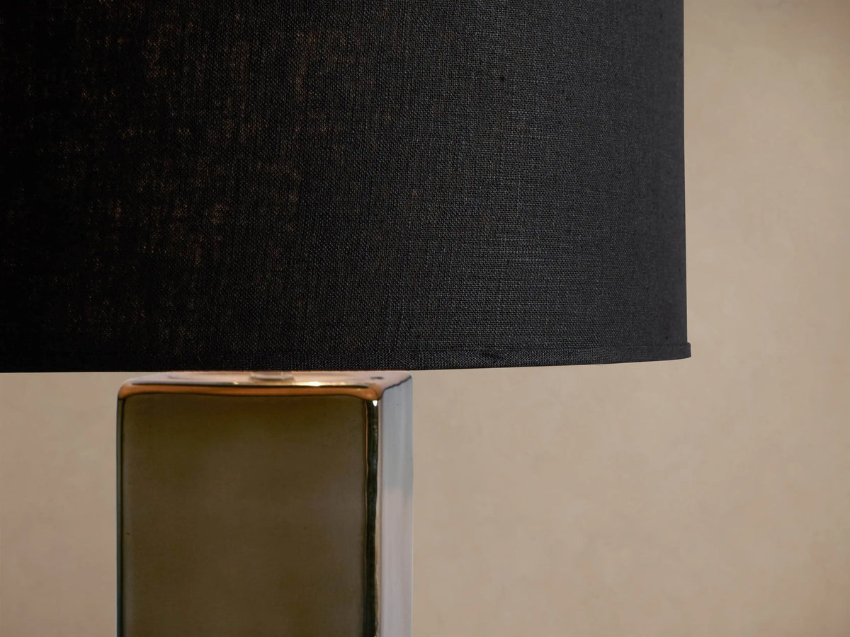 Adrano Table Lamp with a dark linen drum shade on a tall, rectangular cast glass base, gold fading to dark grey, on wood.
