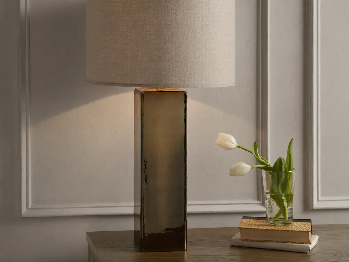 Adrano Table Lamp with antique copper finish base and natural linen drum shade on wooden surface, near books and tulips.