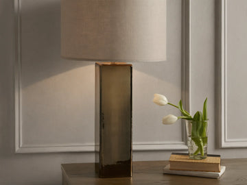 Adrano Table Lamp with antique copper finish base and natural linen drum shade on wooden surface, near books and tulips.