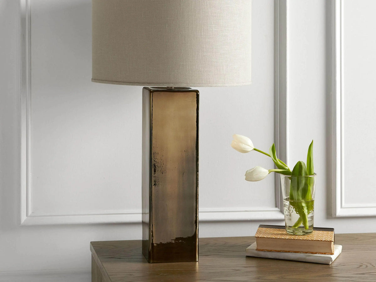 Adrano Table Lamp with antique copper finish base and natural linen drum shade on wooden surface, near books and tulips.