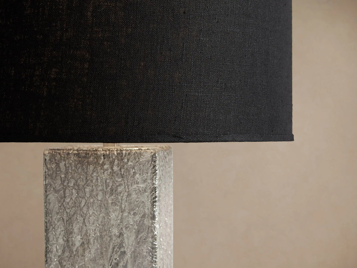 Adrano Table Lamp with a black drum shade and a rectangular, textured silver cast glass base, on a rustic wooden table.