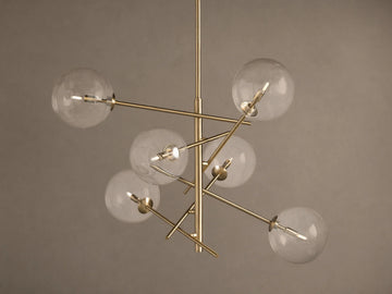 Roscoe Chandelier featuring a brass frame with six clear glass globes extending from a central pole, against a plain wall.