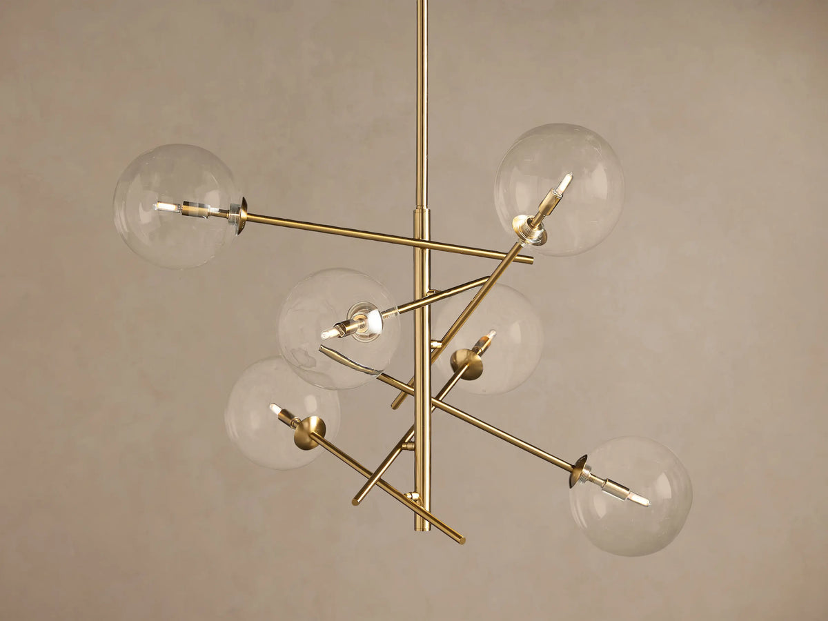 Roscoe Chandelier featuring a brass frame with six clear glass globes extending from a central pole, against a plain wall.