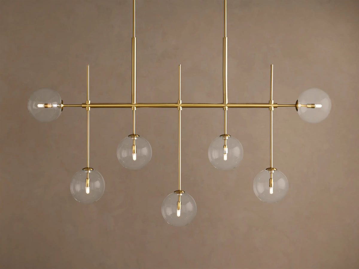 Roscoe Linear Chandelier: horizontal brass frame with seven clear glass globe lights showing a warm glow, against a tan wall.