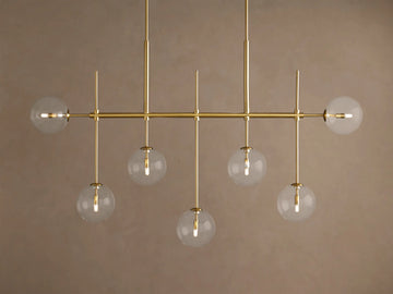 Roscoe Linear Chandelier: horizontal brass frame with seven clear glass globe lights showing a warm glow, against a tan wall.