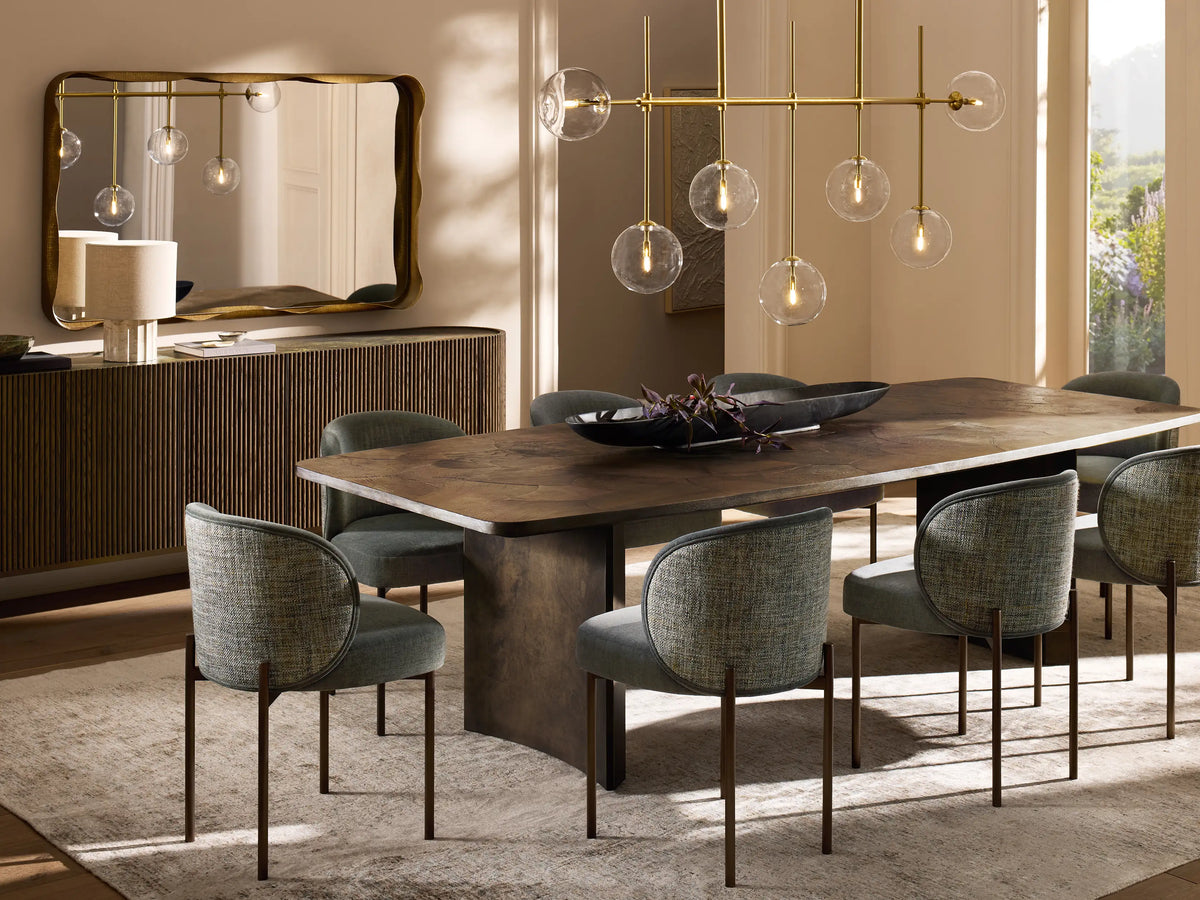 Roscoe Linear Chandelier: horizontal brass frame with seven clear glass globe lights showing a warm glow, against a tan wall.