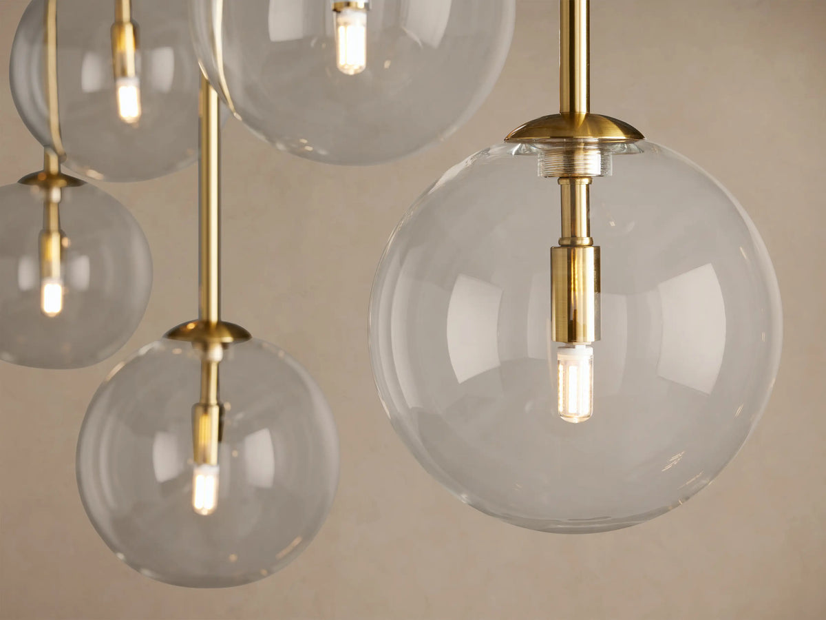 Roscoe Linear Chandelier: Close-up of clear glass spheres, brass-plated rods, and glowing LED bulbs against a light wall.