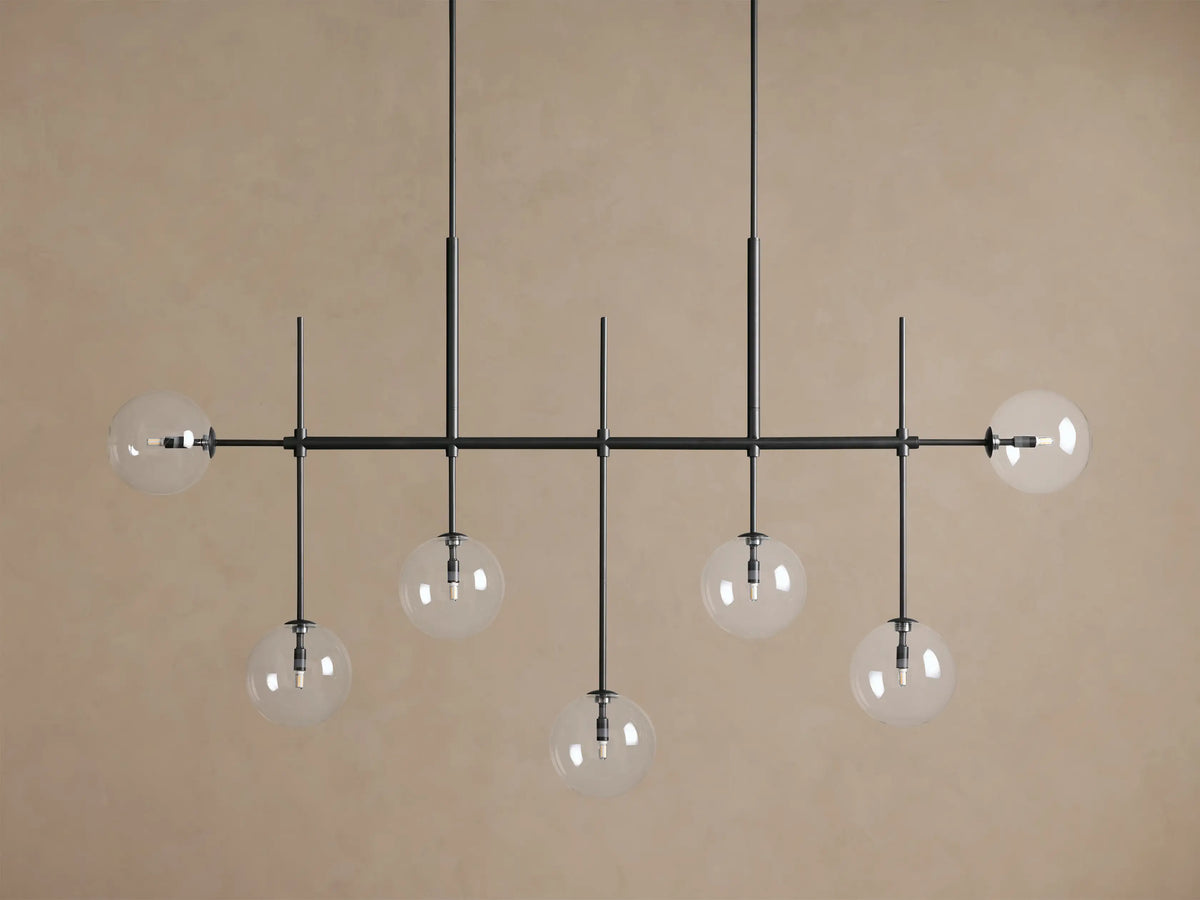 Roscoe Linear Chandelier: black linear frame, 7 clear glass globe lights glowing against a warm brown wall.