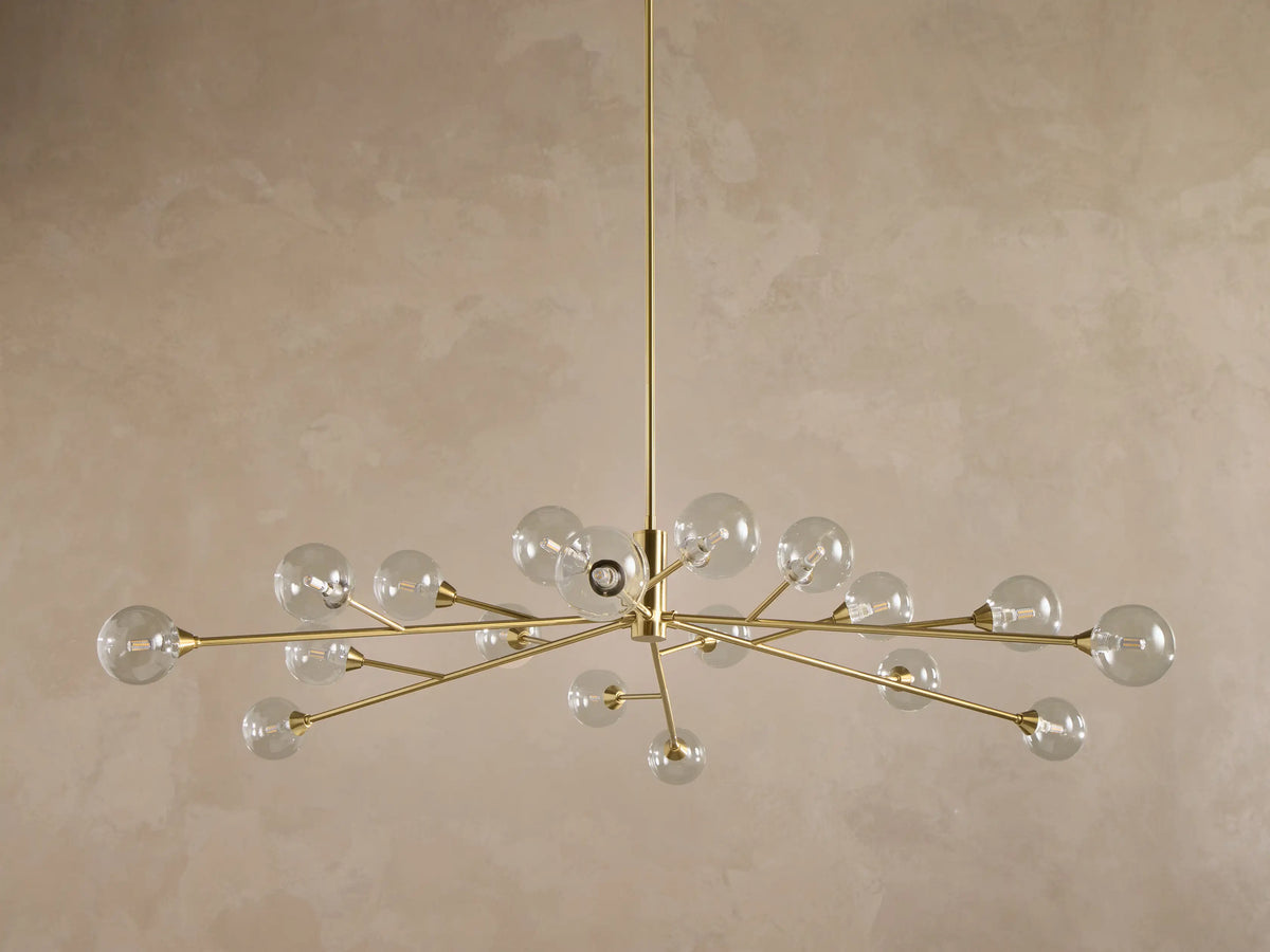 Apollo Round Chandelier: Brass-toned arms with many illuminated clear glass globe bulbs, suspended against a textured wall.