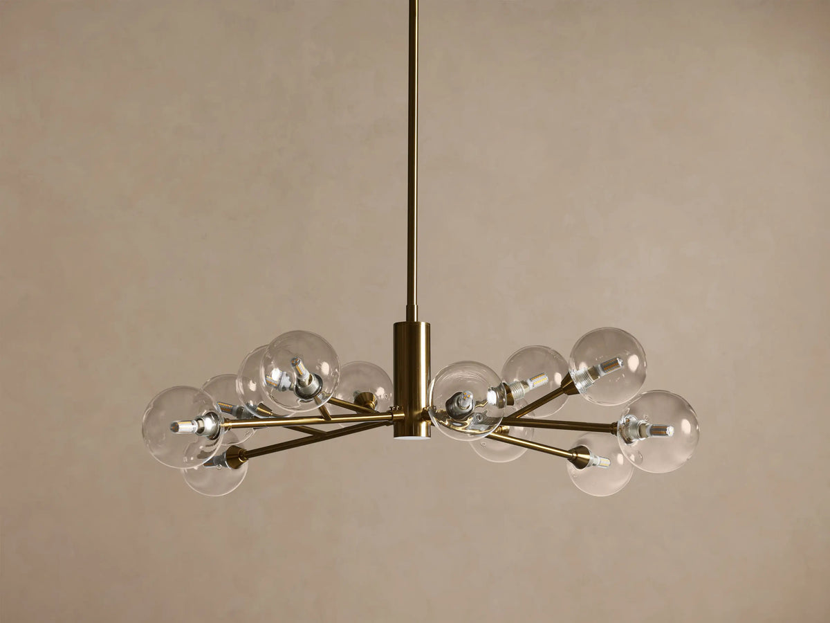 Apollo Round Chandelier, brass frame, clear spherical bulbs glowing brightly, hanging against a warm brown wall.