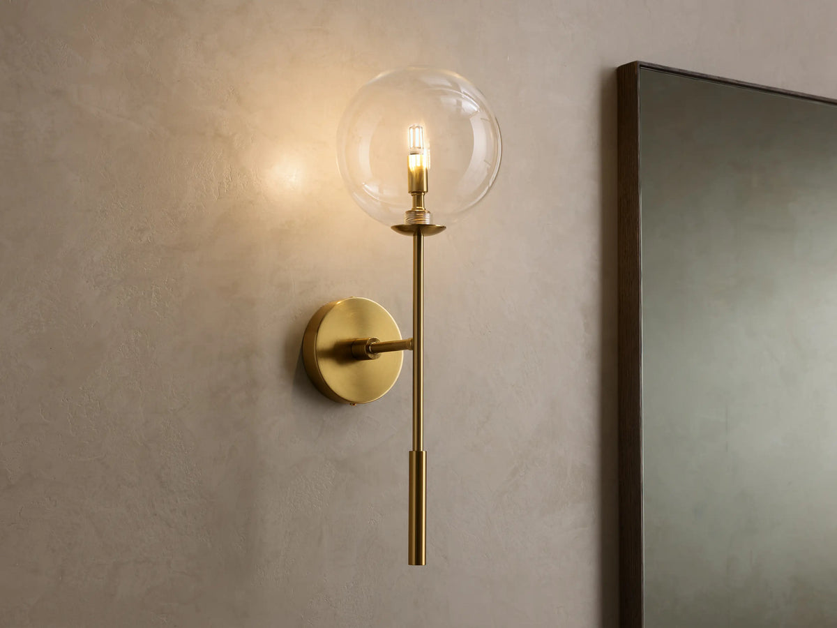 Front view of the brass Roscoe Sconce, featuring a clear glass globe with an illuminated LED bulb, mounted on a wall.