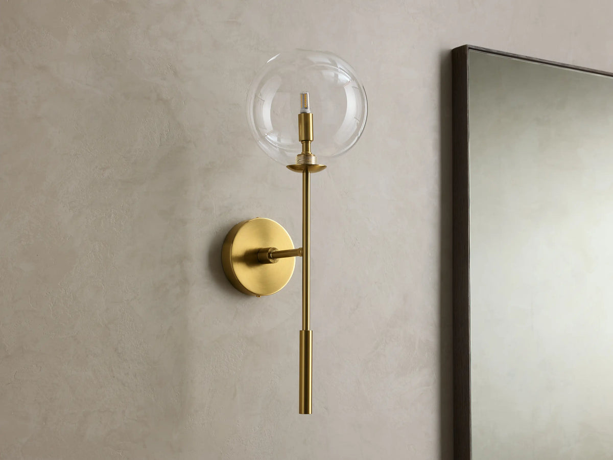 Front view of the brass Roscoe Sconce, featuring a clear glass globe with an illuminated LED bulb, mounted on a wall.