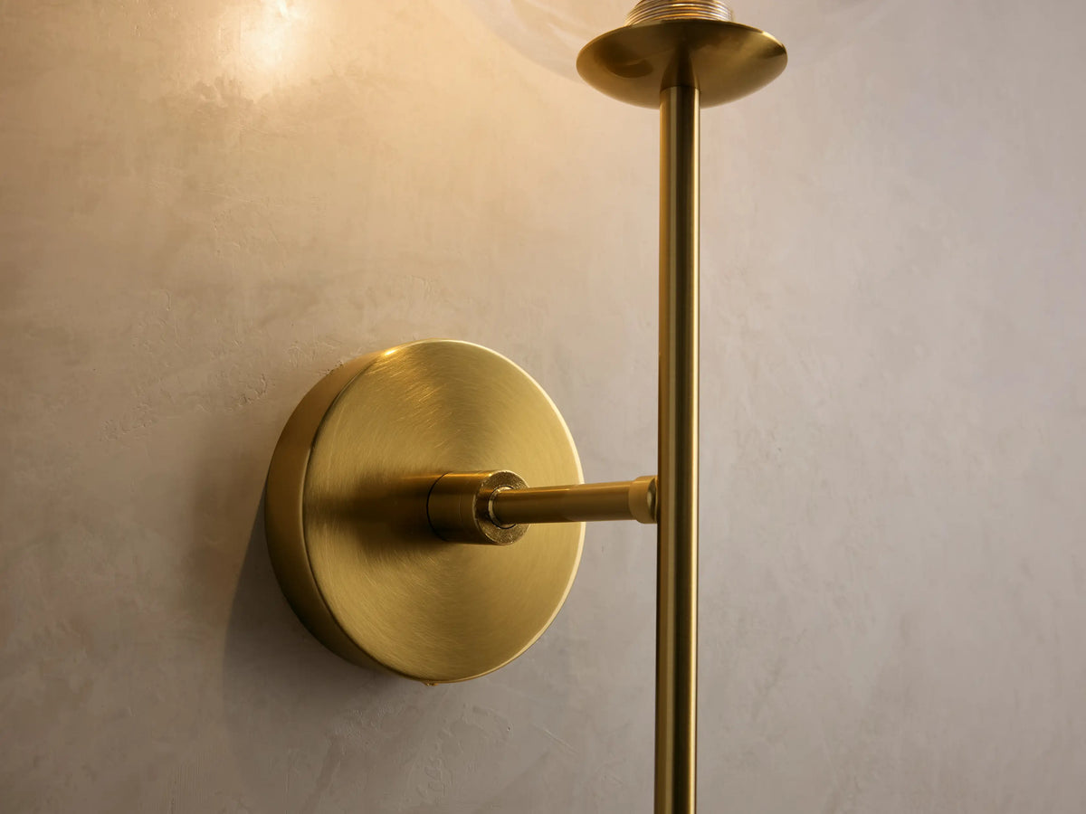 Front view of the brass Roscoe Sconce mounted on a wall, with a clear glass globe and linear rod.