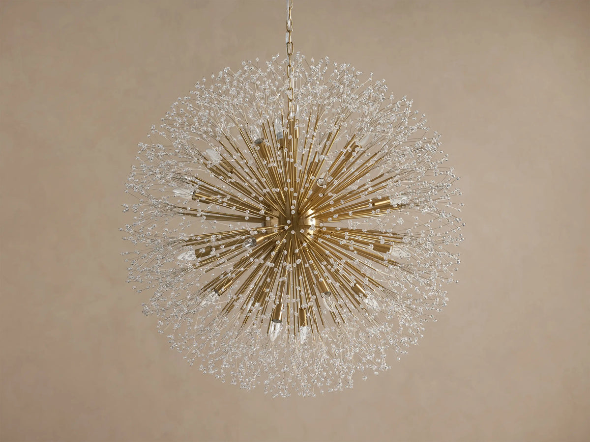 Large, gold-toned Dandelion Round Chandelier with crystal-tipped radiating arms and illuminated bulbs, hanging on a taupe wall.