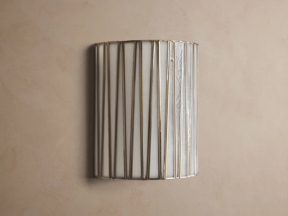 Jeunet Sconce, glowing, with vertical antiqued brass iron strips over translucent white glass, mounted on a neutral wall.