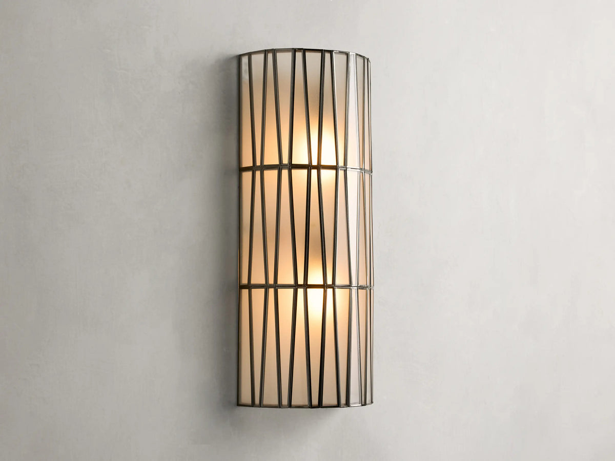 Jeunet Sconce with antiqued brass finish, features two glowing lights behind a cylindrical iron frame with vertical and diagonal lines.