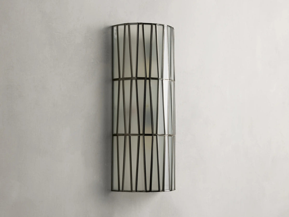 Jeunet Sconce with antiqued brass finish, features two glowing lights behind a cylindrical iron frame with vertical and diagonal lines.