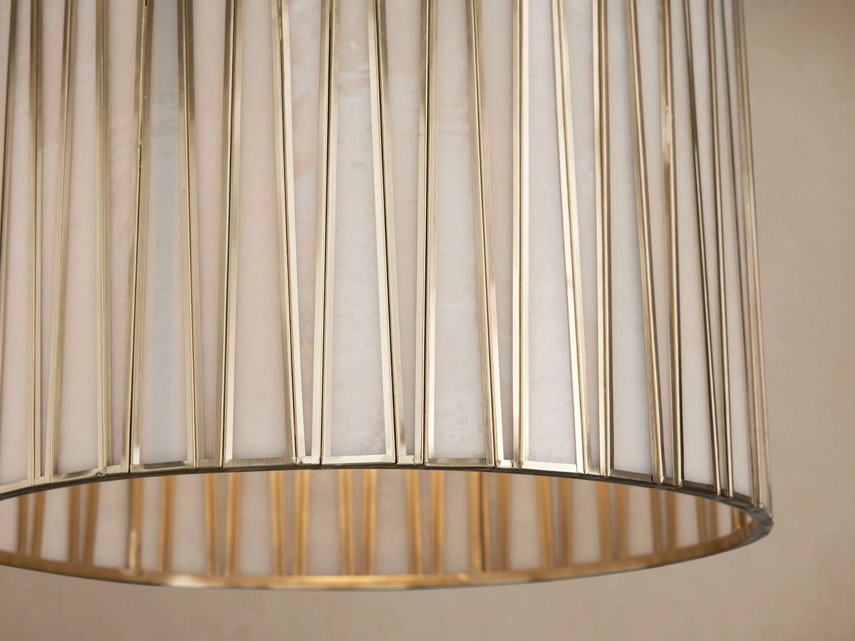 Jeunet Pendant, cylindrical with vertical antiqued brass metallic bars over a light inner shade, hanging against a beige wall.
