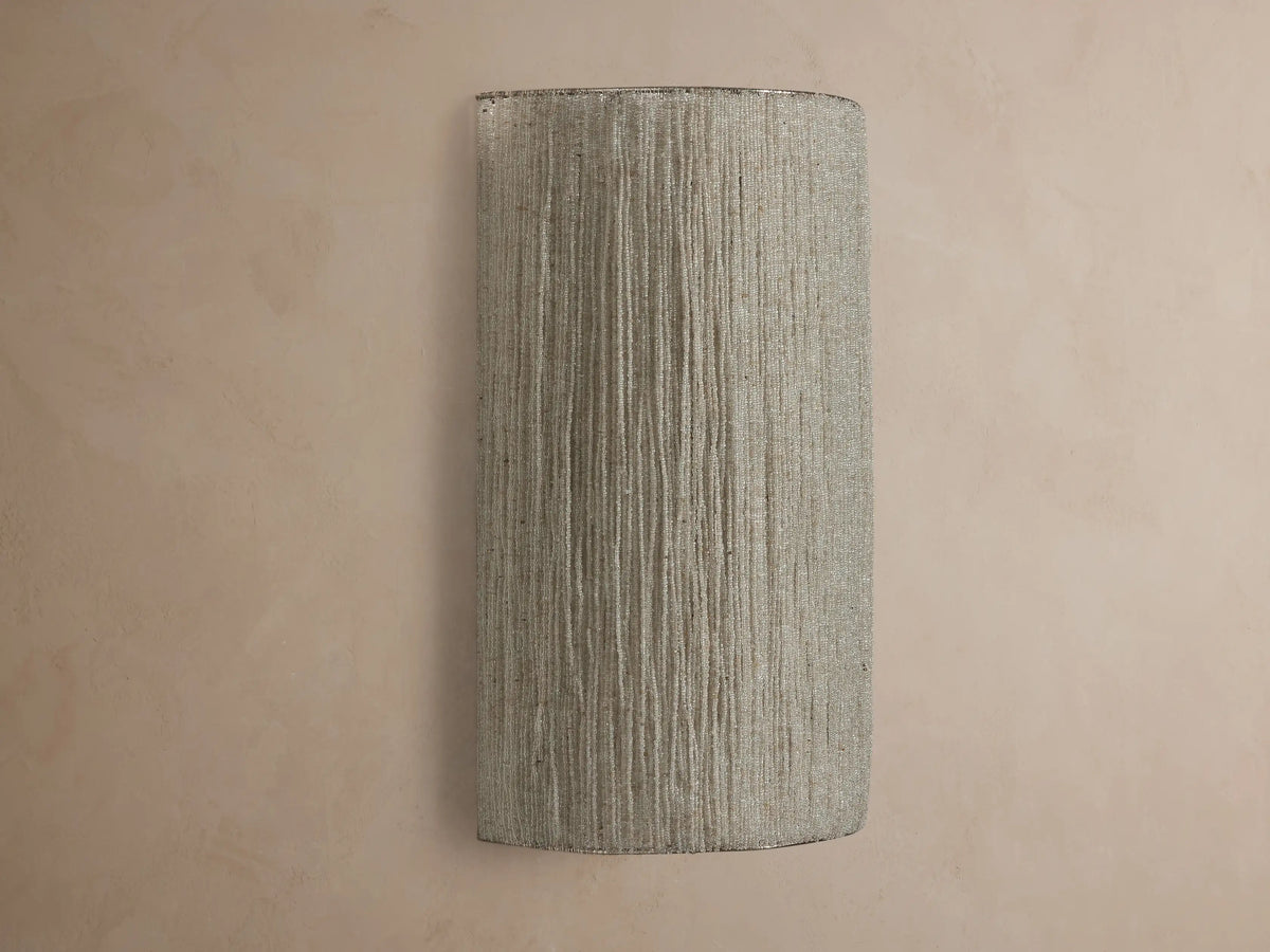 Romilly Beaded Sconce, vertical, elongated, warm light shining through its textured, light-colored strung glass beads on a neutral wall.