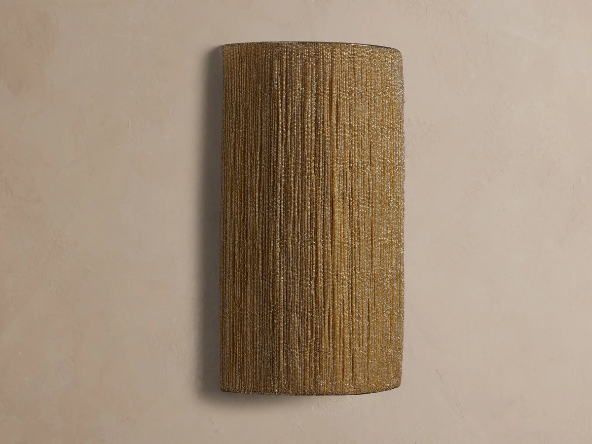 Romilly Beaded Sconce, rectangular & curved, with golden glass beads. Illuminated from within, casting a warm glow on a wall.