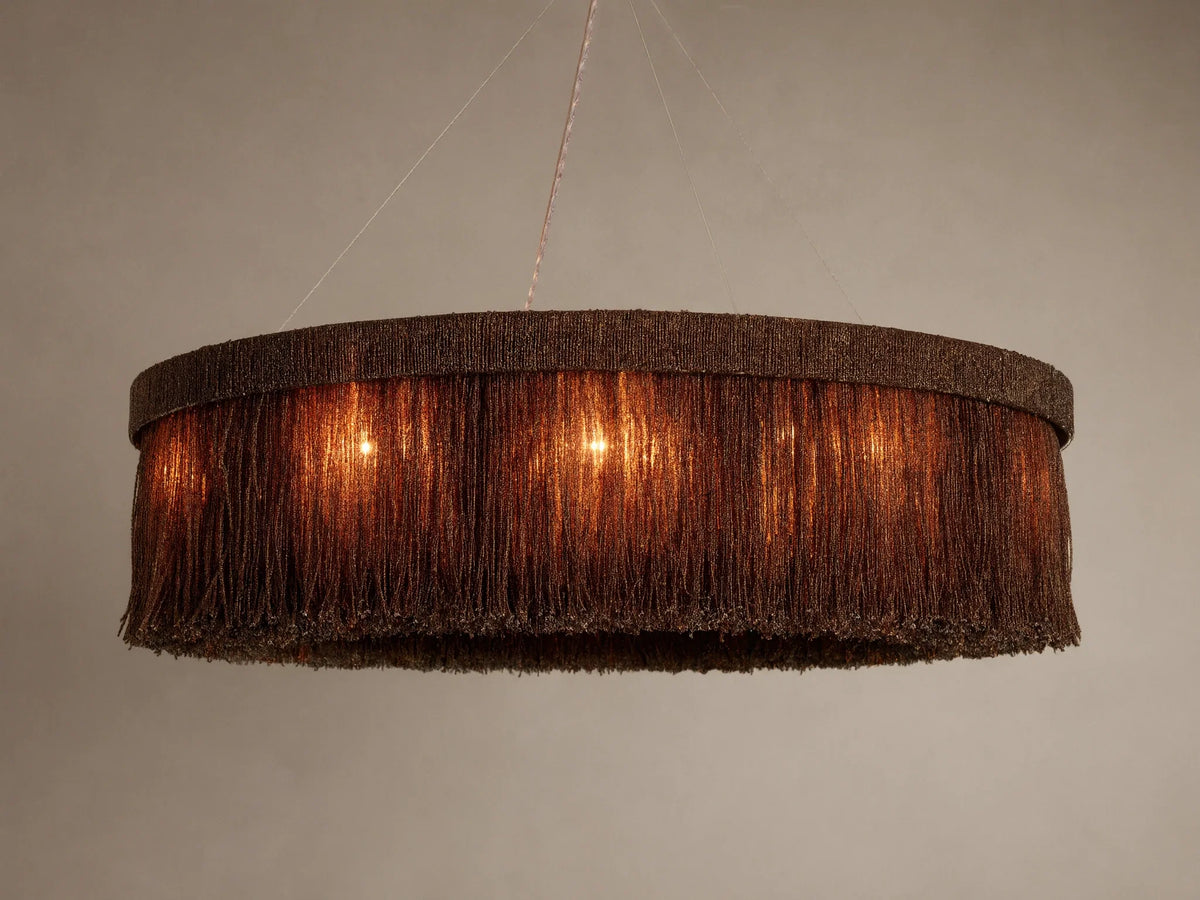 Romilly Round Chandelier, dark bronze beaded fringe, emitting a warm orange glow from internal lights, hanging against light wall.