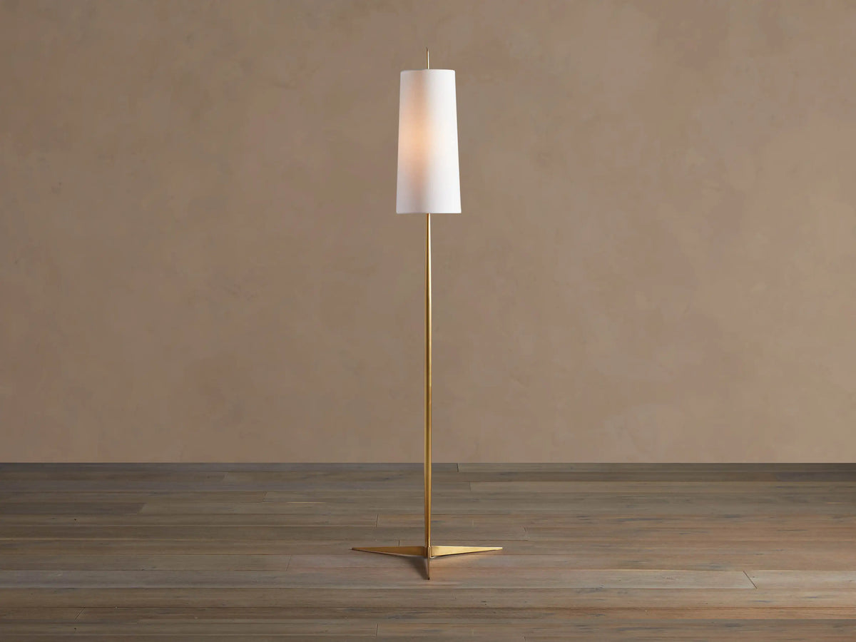 Lancia Floor Lamp with slender brass pole, triangular base, and tall, glowing white linen shade on a wood floor.