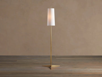 Lancia Floor Lamp with slender brass pole, triangular base, and tall, glowing white linen shade on a wood floor.