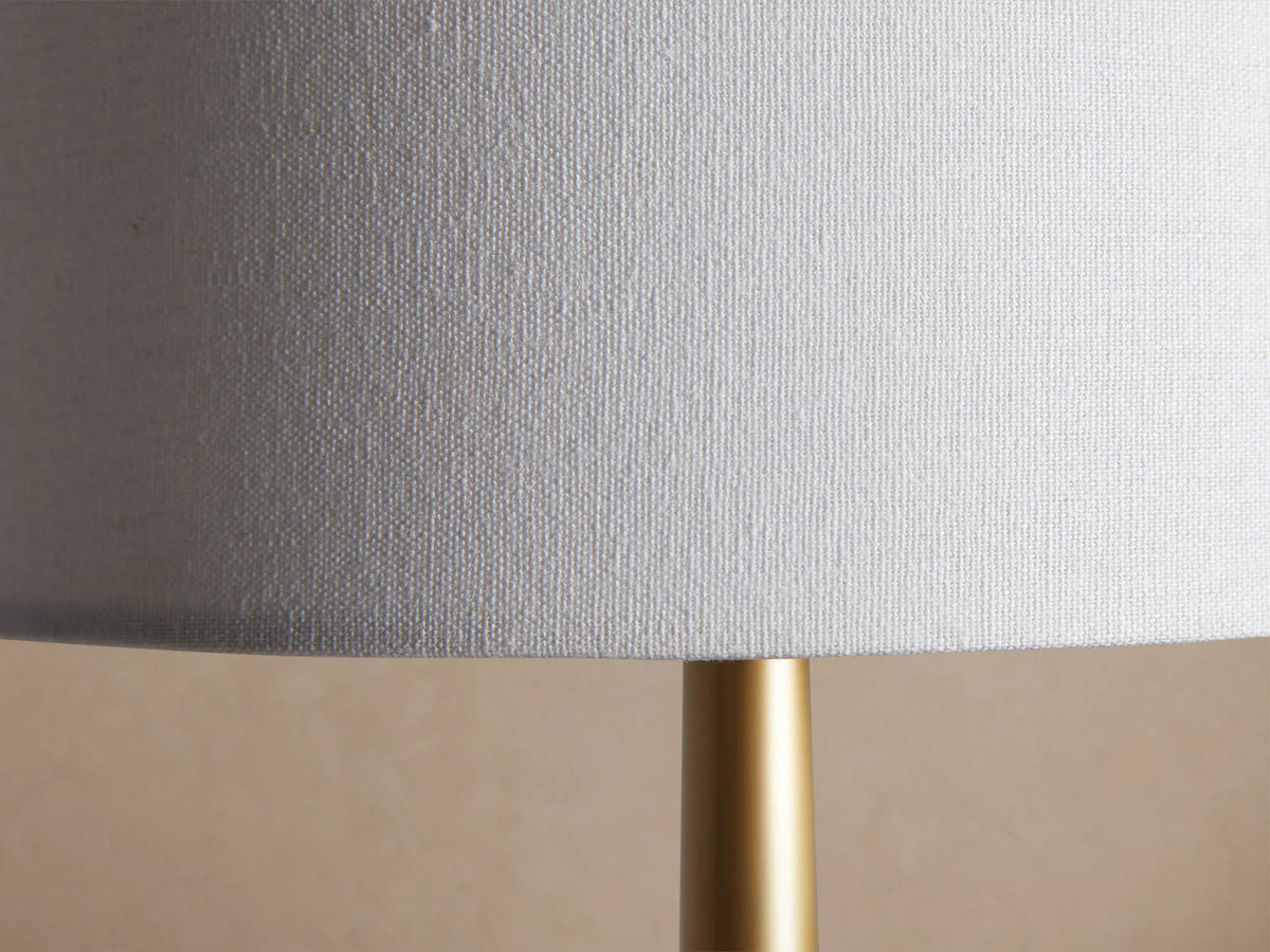 Lancia Floor Lamp: tall, slender brass-finished lamp with a white cylindrical linen shade, standing upright on a wood floor.