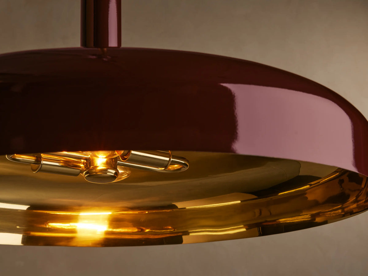 A burgundy pendant light with a glossy finish, featuring a cylindrical stem and a brass rod, set against a beige wall.