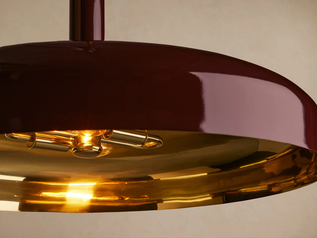 A maroon pendant light with a glossy finish hangs from a brass rod against a beige background, featuring a flat, wide shade.