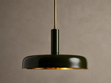 Bonham Pendant: Dark green dome-shaped light with a brass interior hangs from a brass rod against a textured warm grey wall.