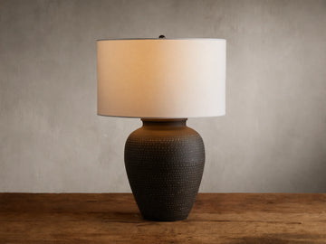 Howell Table Lamp with an embossed, dark powder-coated base and a wide, white cotton linen shade, on a wooden surface.