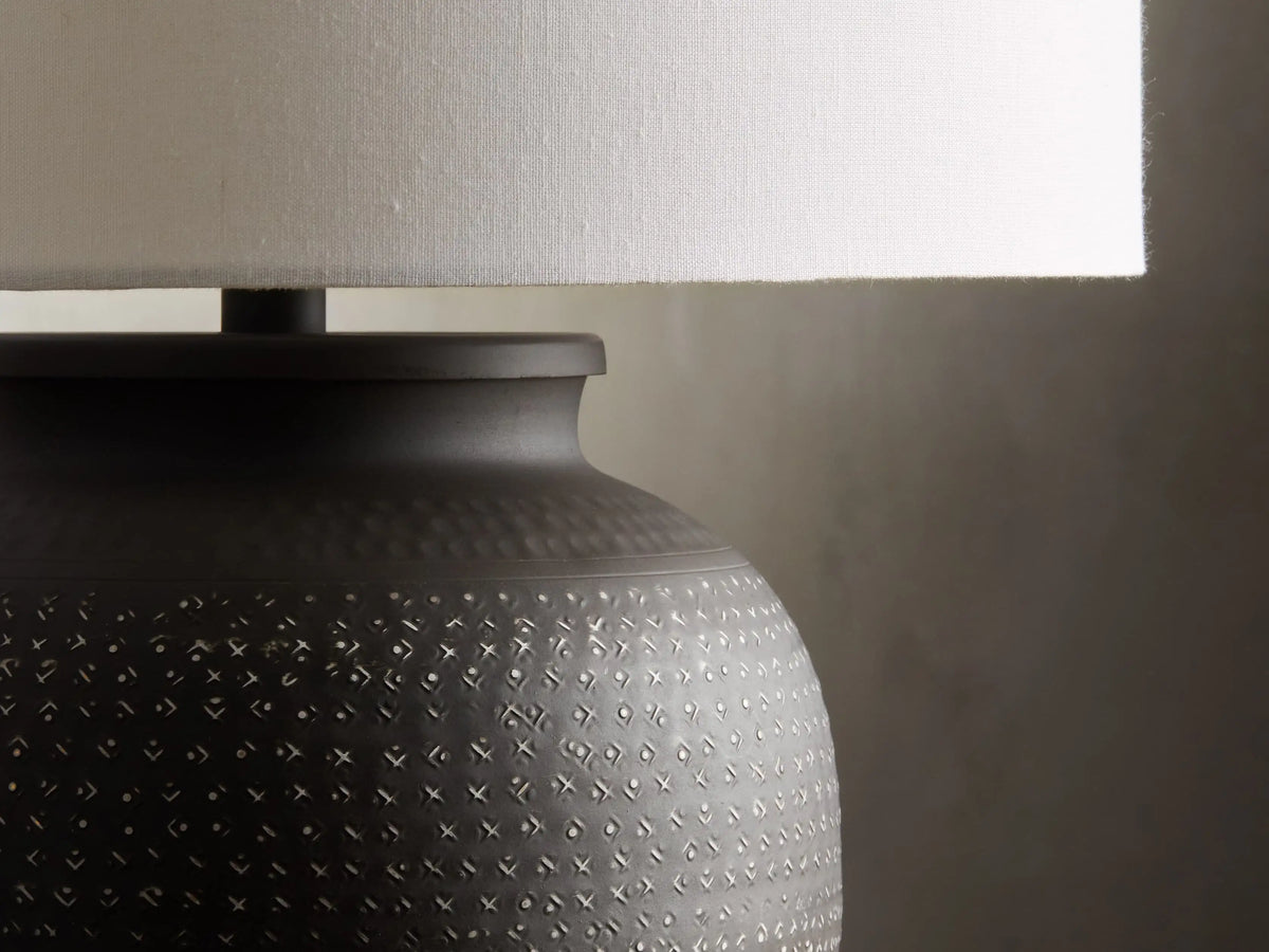 Howell Table Lamp with an embossed, dark powder-coated base and a wide, white cotton linen shade, on a wooden surface.