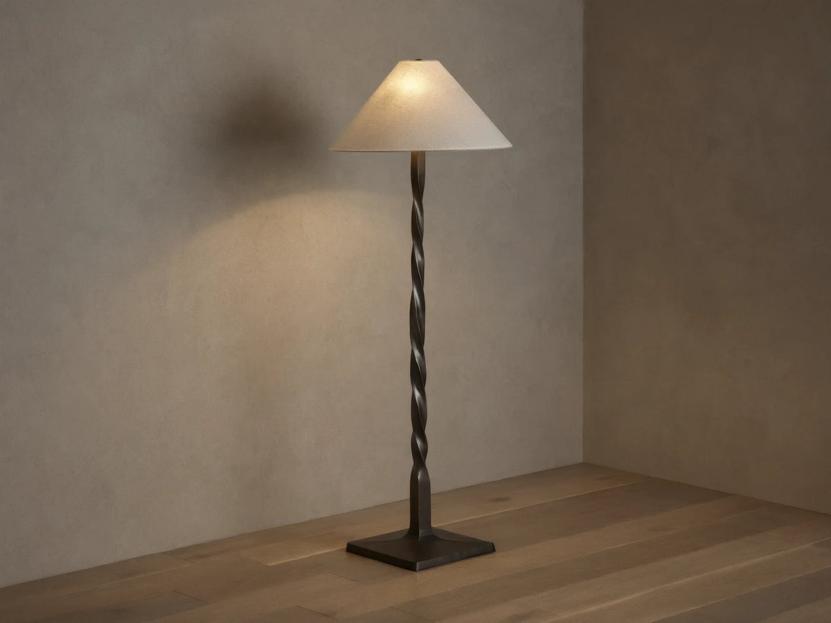 A tall floor lamp with a twisted bronze base and a conical cream lampshade, placed on a wooden floor against a neutral wall.