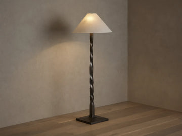 A tall floor lamp with a twisted bronze base and a conical cream lampshade, placed on a wooden floor against a neutral wall.