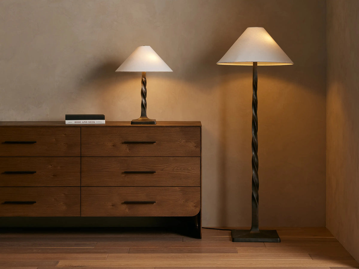A tall floor lamp with a twisted bronze base and a conical cream lampshade, placed on a wooden floor against a neutral wall.