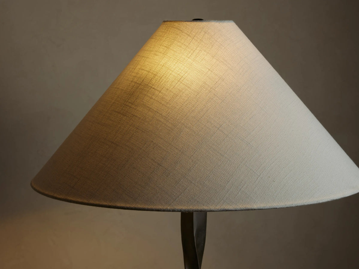 A floor lamp with a twisted dark metal base and a wide, white conical shade, positioned against a beige wall on wooden flooring.