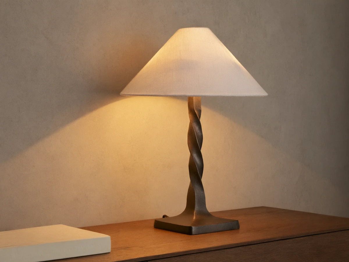 A table lamp with a twisted bronze base and a beige, conical fabric shade, casting a warm glow on a wooden surface.