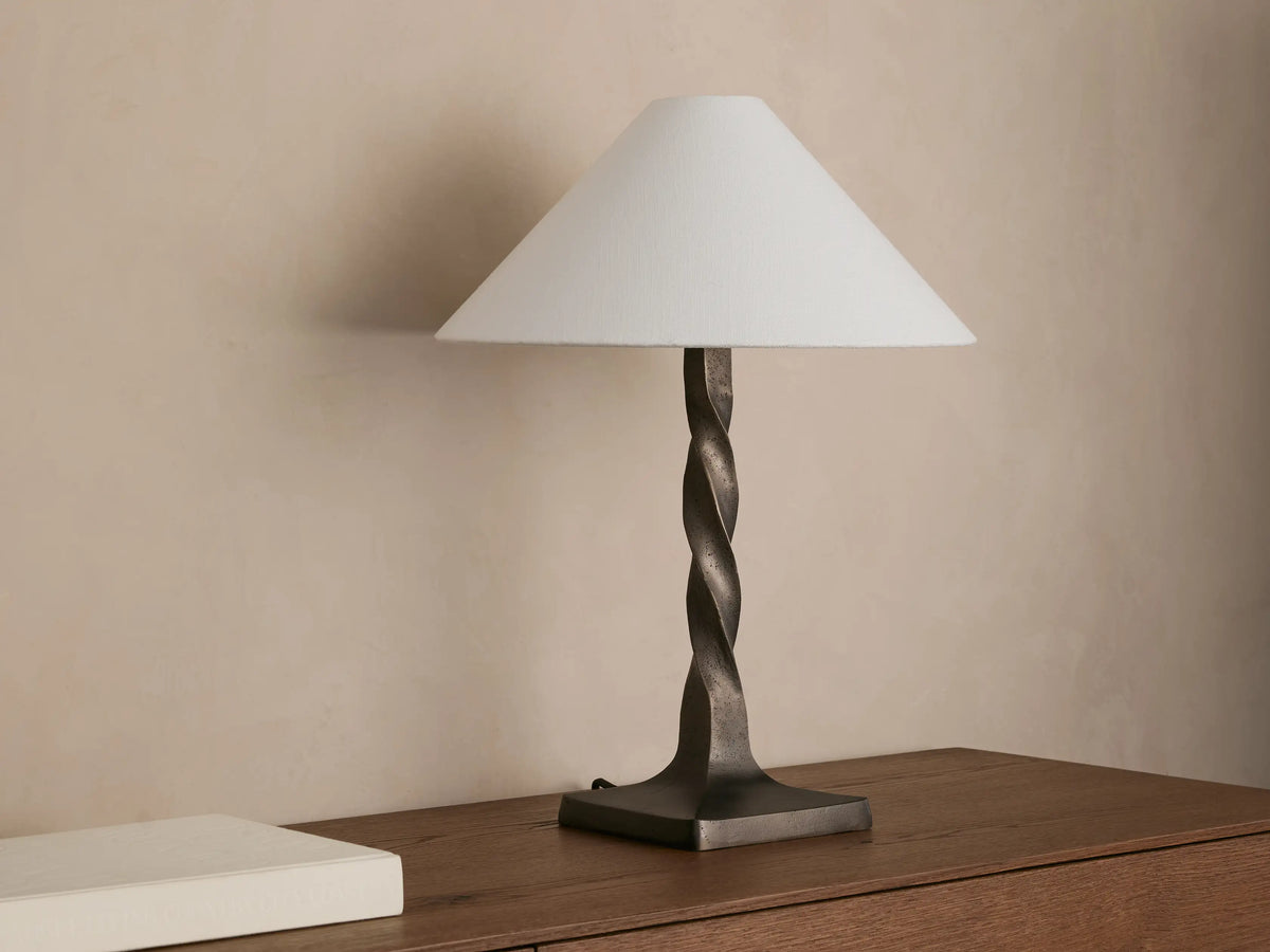 A table lamp with a twisted bronze base and a beige, conical fabric shade, casting a warm glow on a wooden surface.