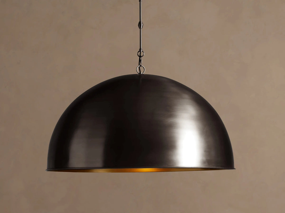 Watley Dome Pendant. Dark oxidized brass dome light, showing a gleaming golden interior, hanging against a brown wall.