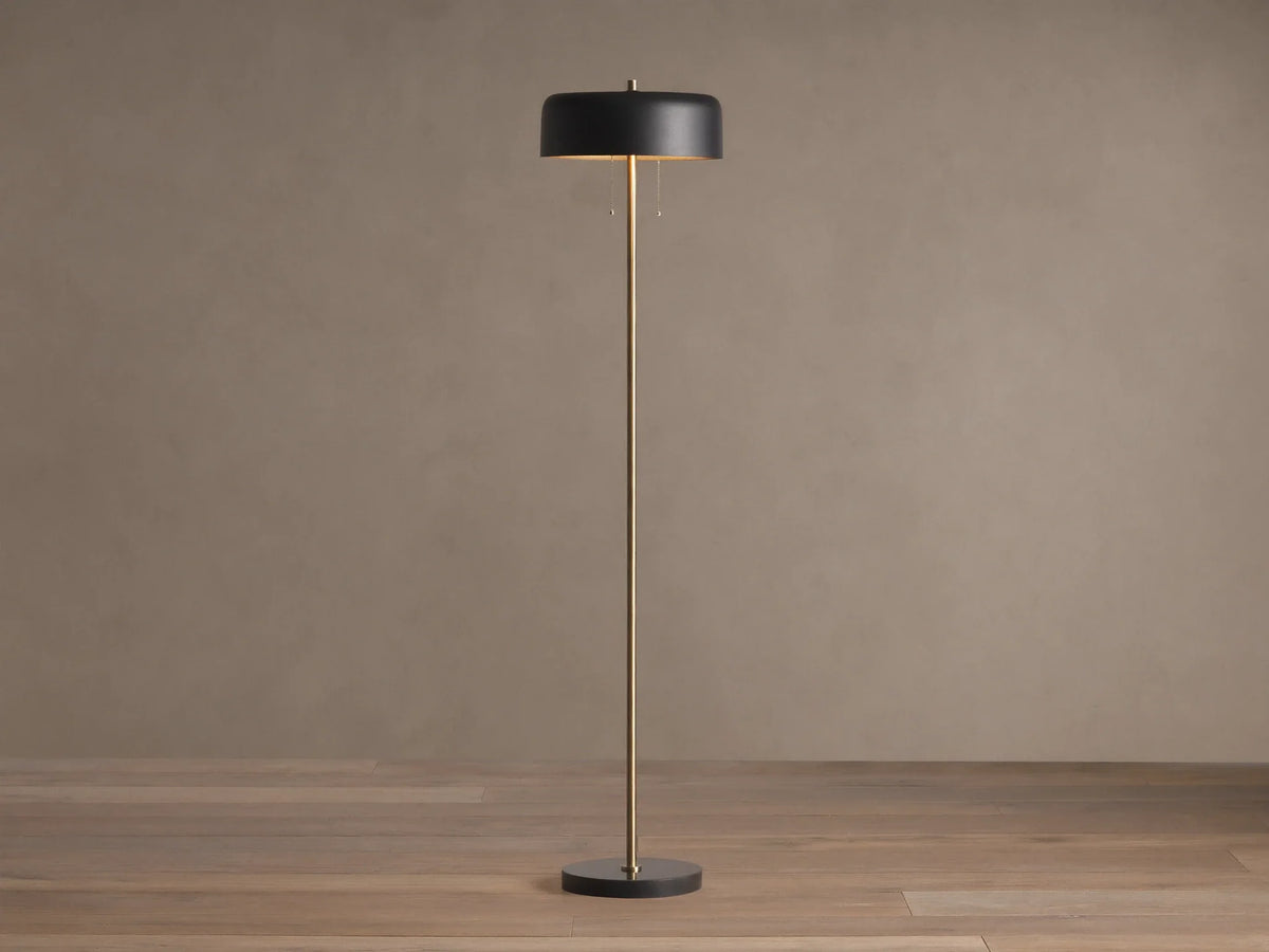Oliver Floor Lamp: upright brass stem, black cylindrical shade with warm downward glow, black circular base on wooden floor.