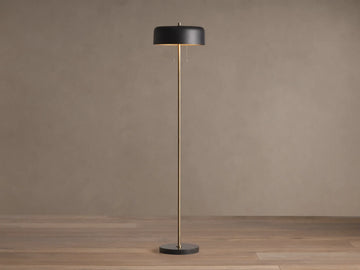 Oliver Floor Lamp: upright brass stem, black cylindrical shade with warm downward glow, black circular base on wooden floor.