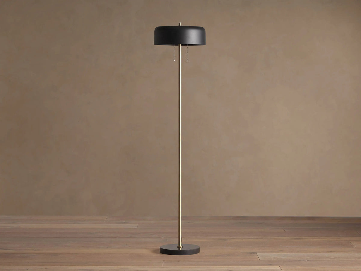 Oliver Floor Lamp: upright brass stem, black cylindrical shade with warm downward glow, black circular base on wooden floor.