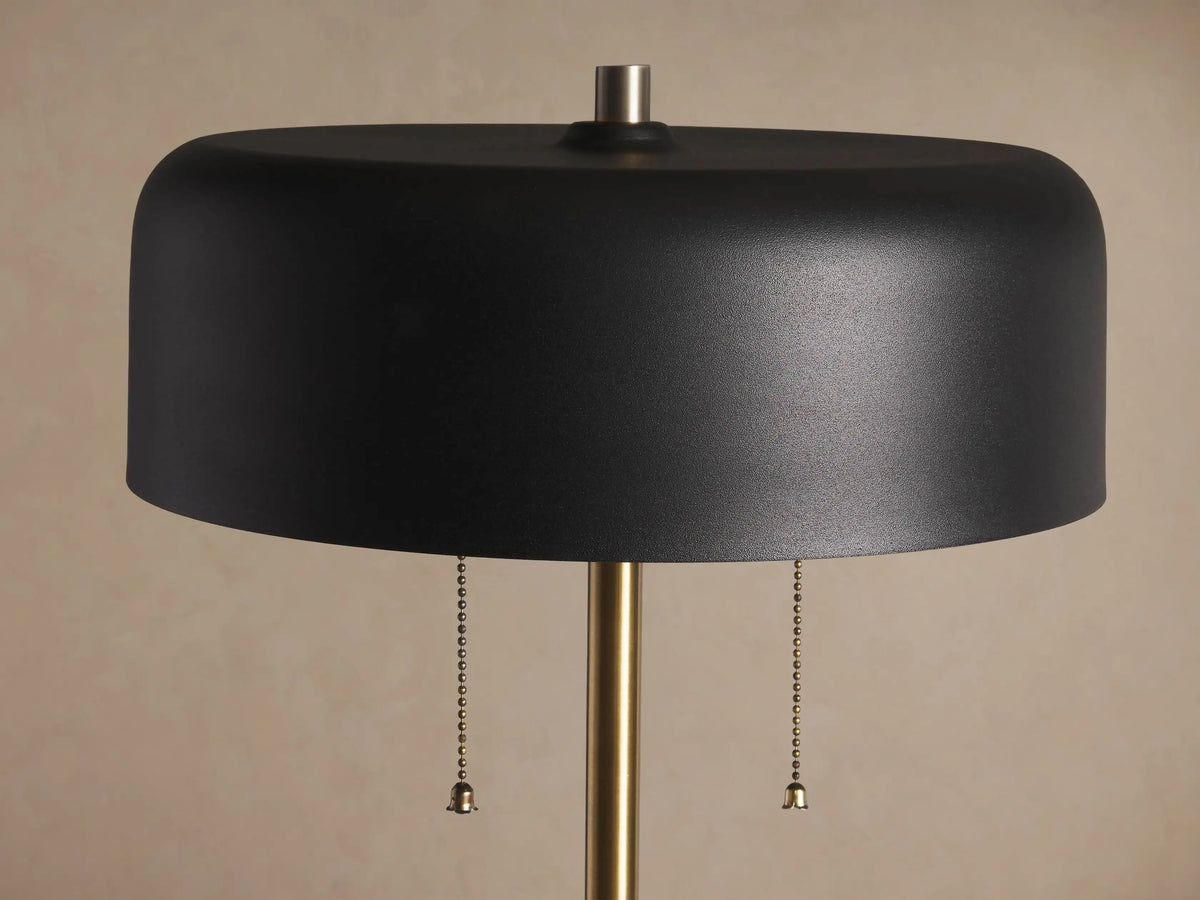Oliver Floor Lamp with a slender brass-finished pole, black cylindrical shade, and circular base on a wooden floor.